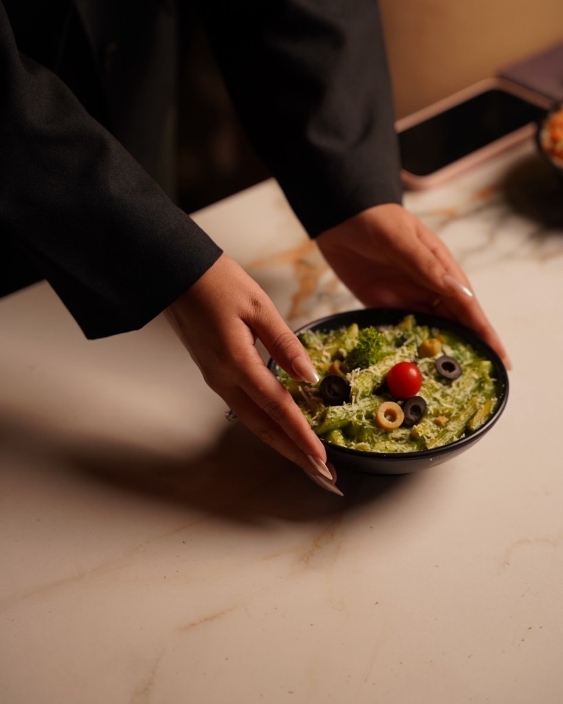 A bowl of green pasta with olive, tomato, and cheese